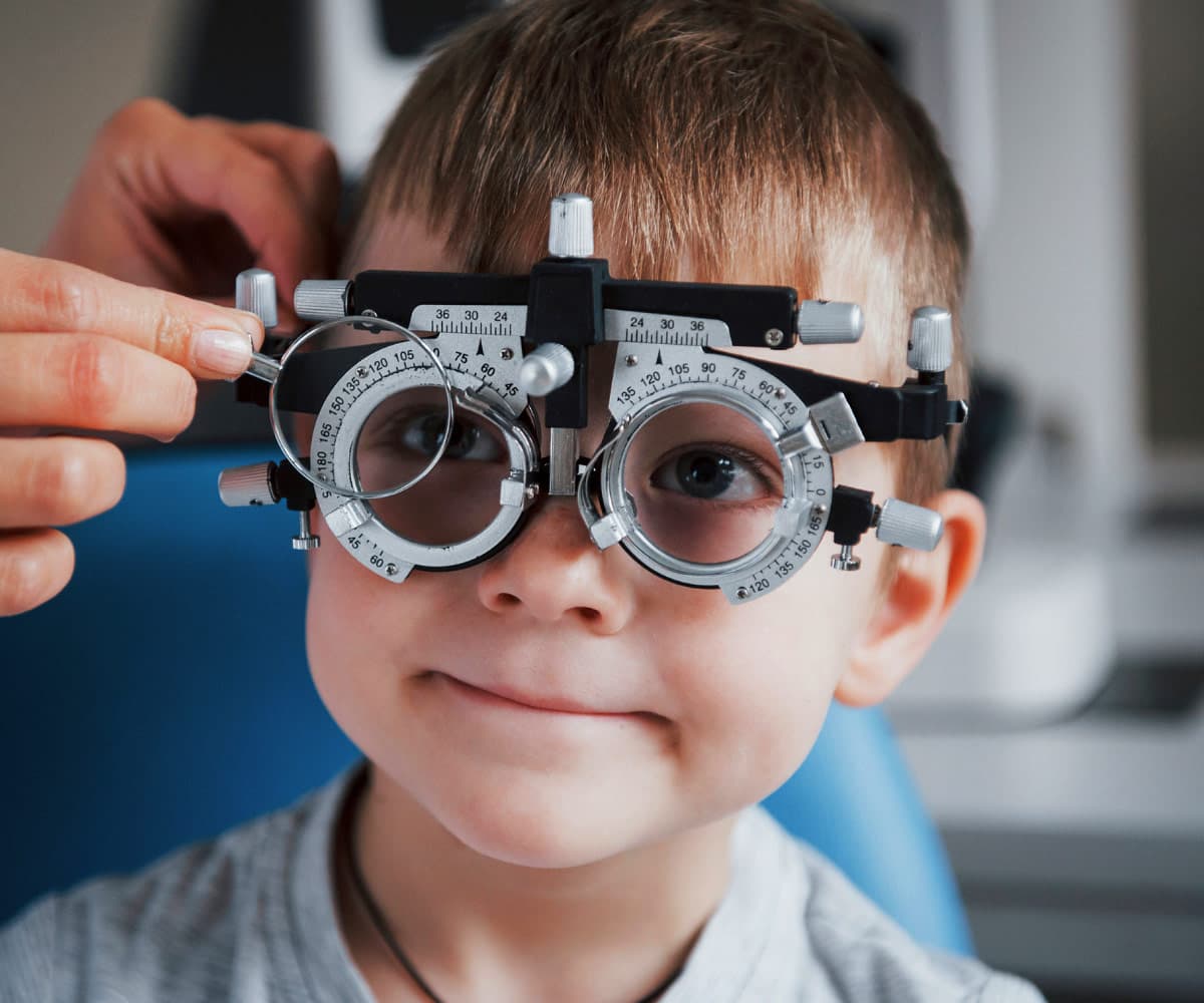 child-eye-test A young child sits in a vision testing chair wearing trial frames for an eye exam. An adults hands adjust the frames to fit properly. The child looks ahead with a slight smile.