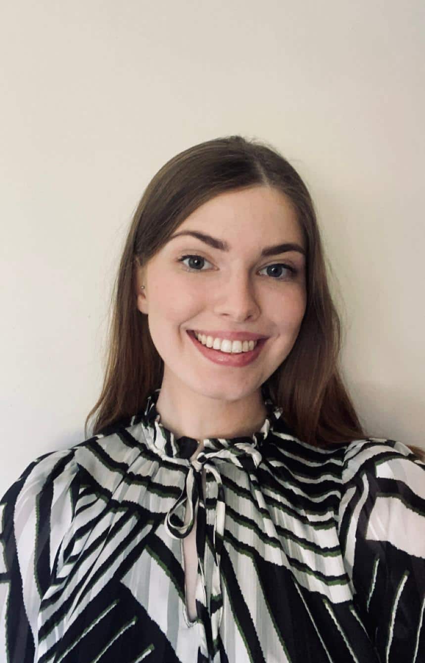 Aoife Lynch (1) Aoife Lynch Optometric Advisor to Optometry Ireland person with long brown hair smiles at the camera. They are wearing a black and white patterned top with a bow at the neck. The background is plain white.