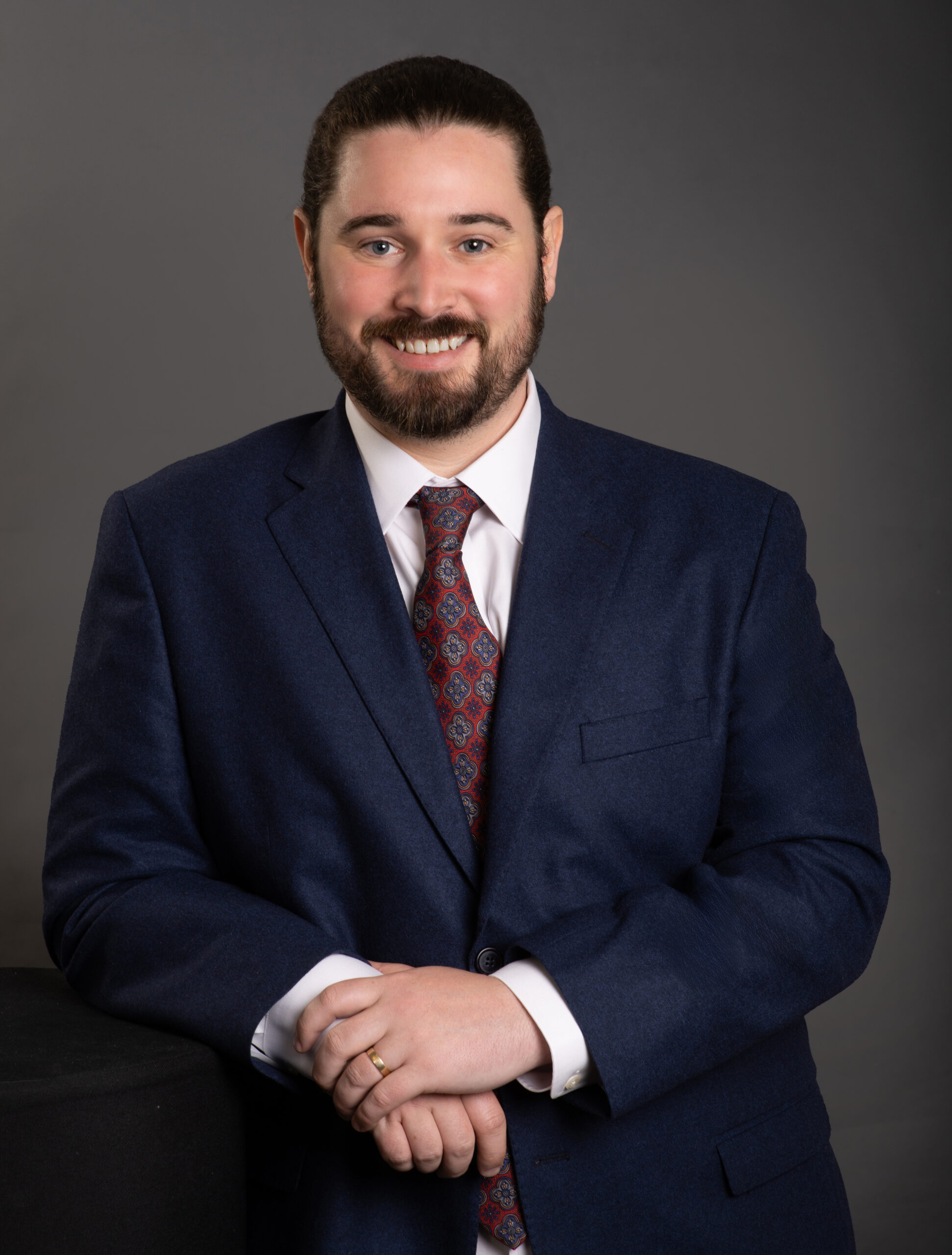 Smiling man with beard and hair tied back in a navy suit, white collar shirt with patterrned wine tie with one hand causally resting on the other in front of his body